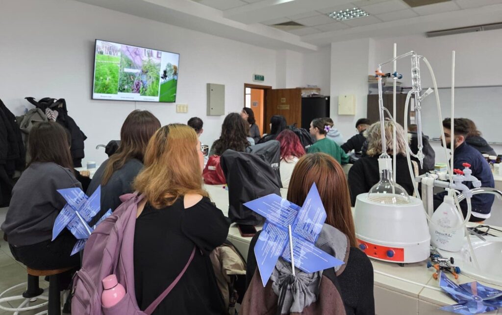 Elevi văzuți din spate într-un laborator de chimie, cu moriști de hârtie personalizate cu materialele proiectului și aparatură de laborator în prim-plan.
