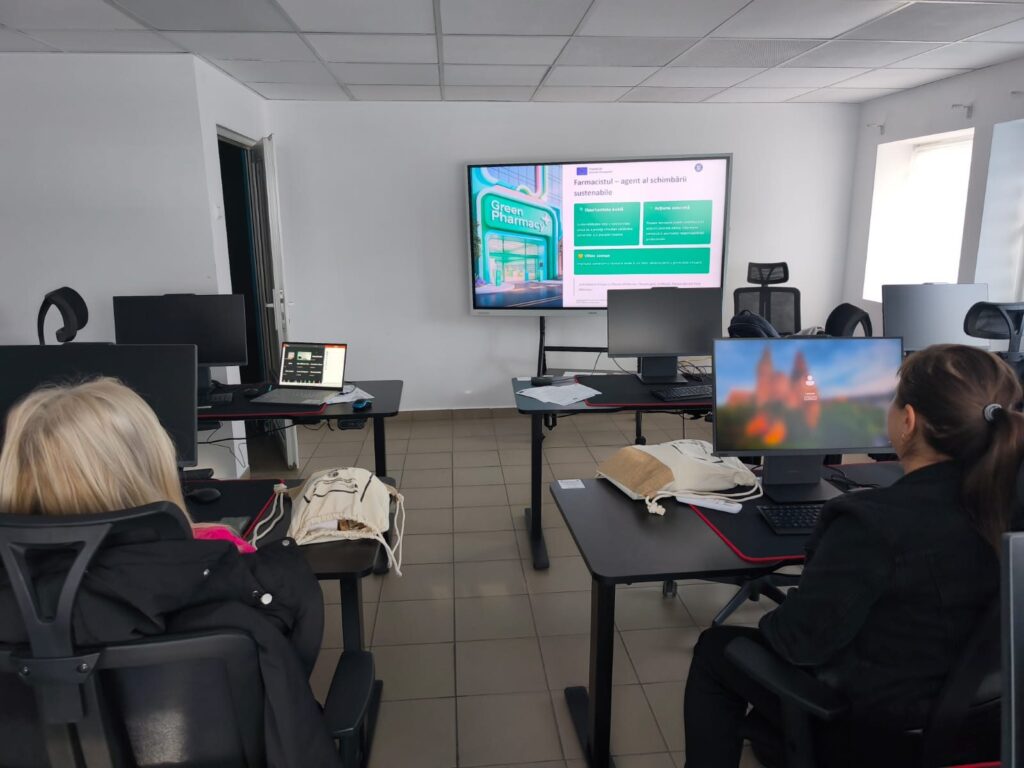 Cursanți așezați la posturi de lucru, văzuți din spate, urmărind un ecran pe care este proiectat un slide al proiectului cu emblema Uniunii Europene.