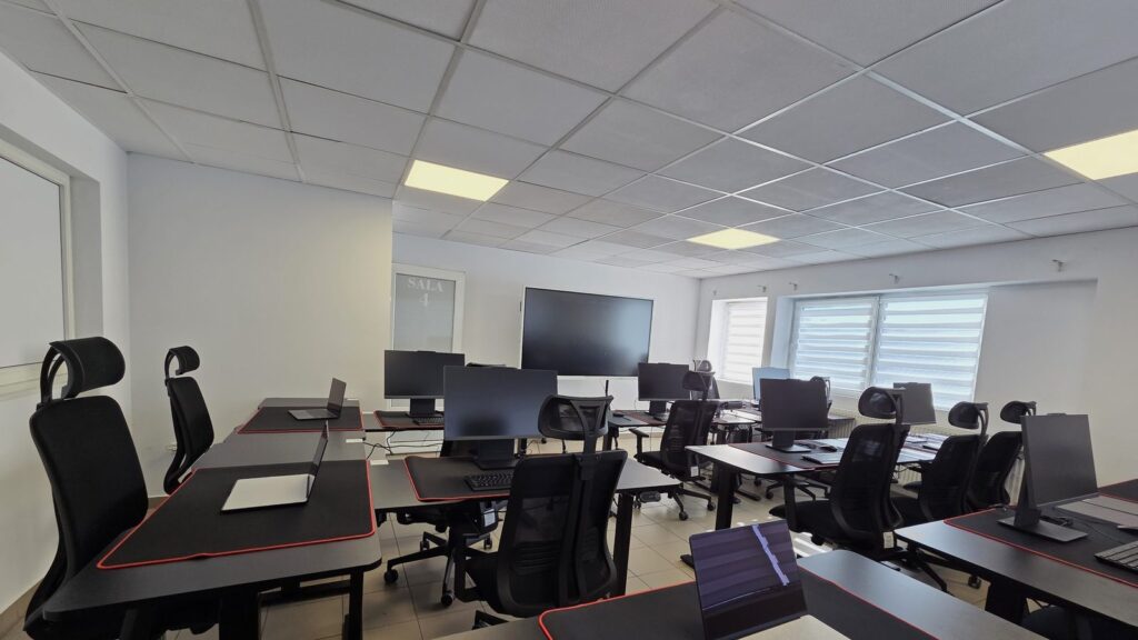 Sală de formare cu douăsprezece posturi de lucru echipate cu monitoare și scaune ergonomice, pregătită pentru cursurile de competențe digitale.