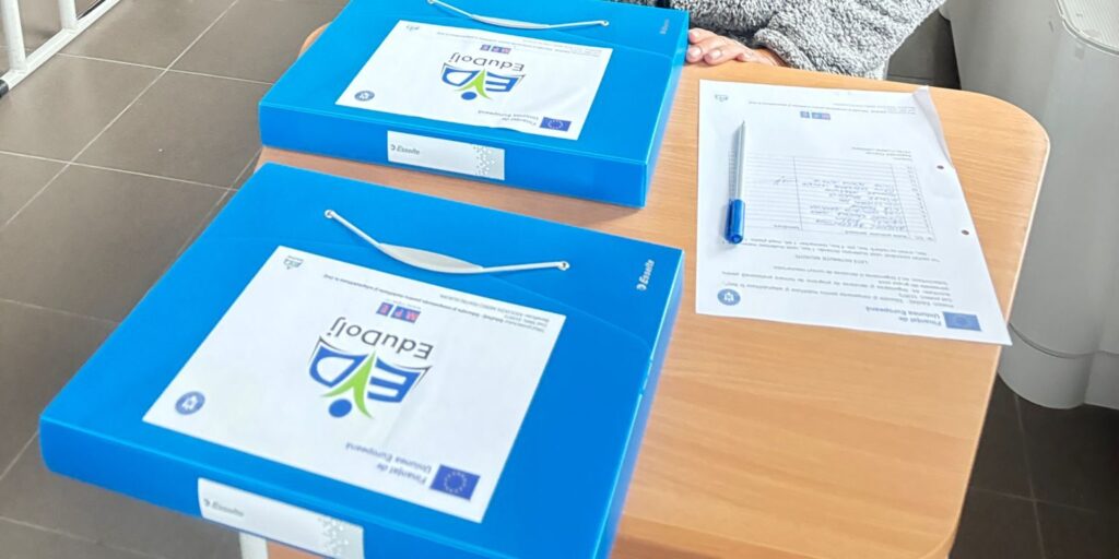 Două mape albastre cu însemnele proiectului EduDolj și ale Uniunii Europene, alături de o fișă de lucru și un pix, pe o bancă de școală.